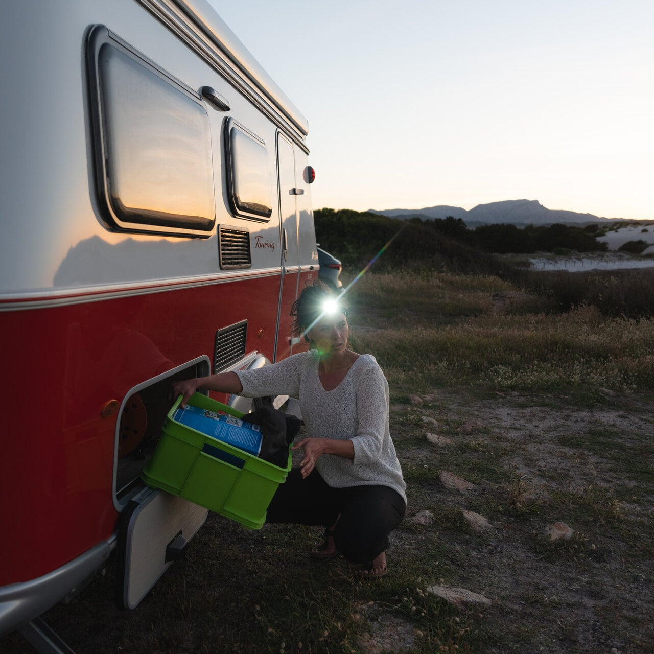 Person kneeling next to a red and white camper van with a light on, in a natural setting.