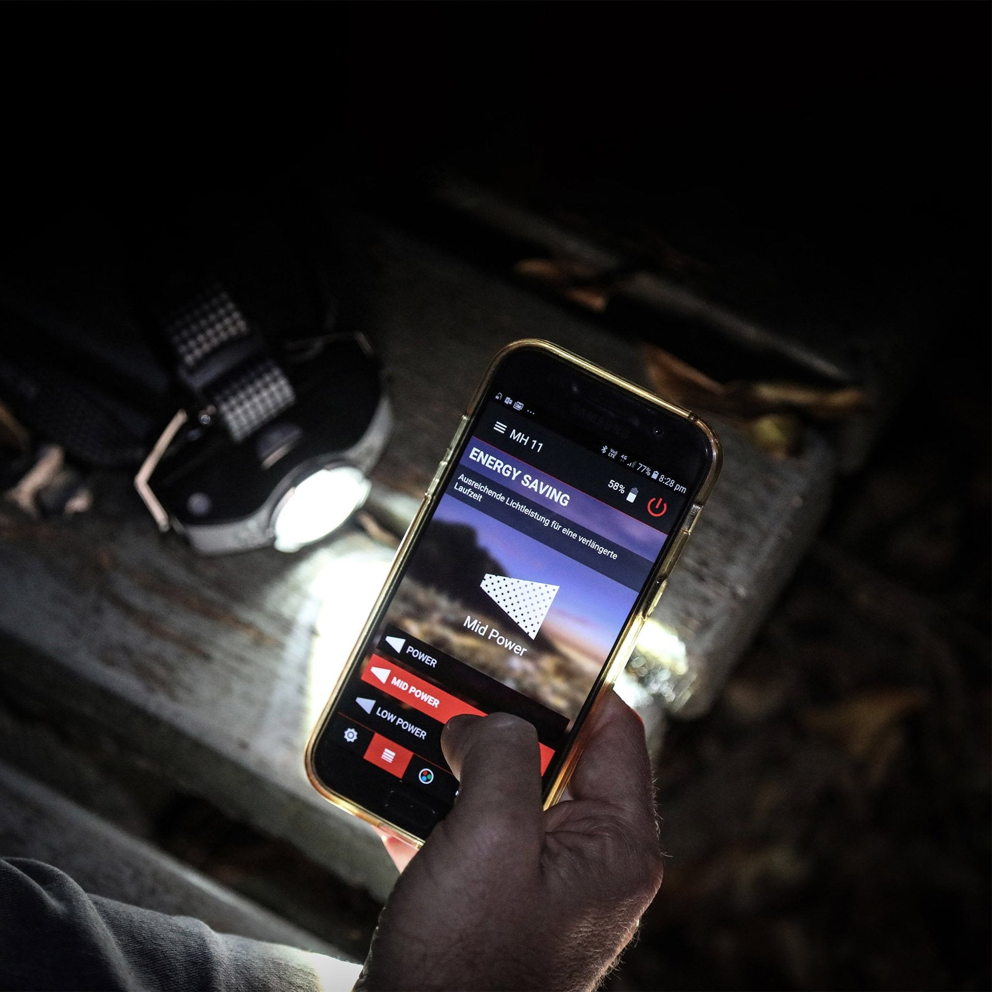 Hand holding a smartphone with an app interface in a dimly lit room, with a flashlight on a surface in the background.